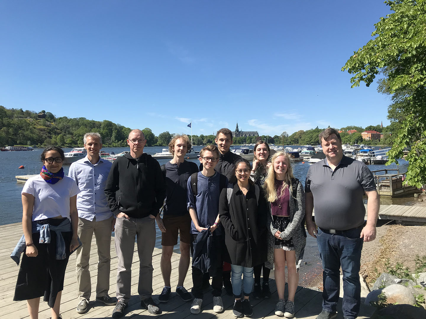 A group of students and three people from the company Emvico, posing for the photograph. It's summertime and in the background you can see water, some boat docks and several boats.