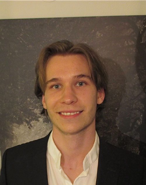 Image of a young man smiling, standing in front of a wall, wearing a dark suit jacket and white shirt.