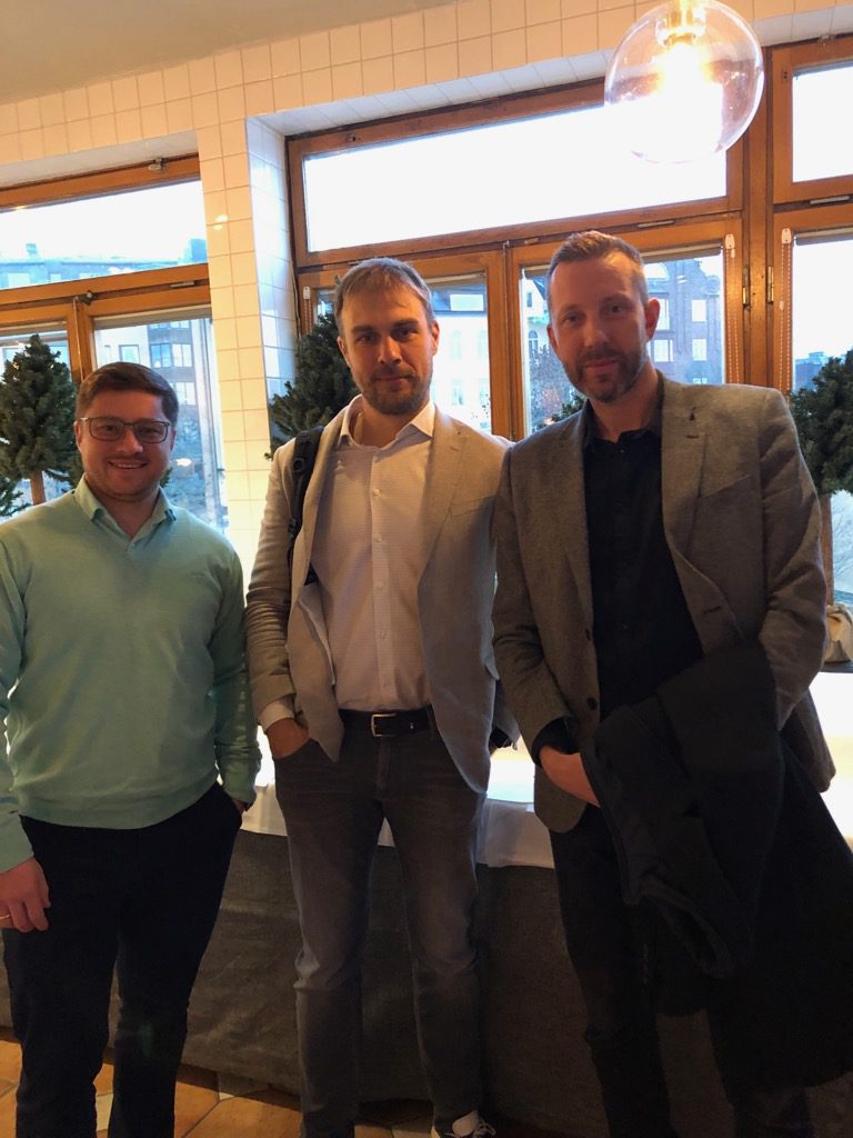 Three men posing for a the picture. They are inside an office, standing in front of some windows.